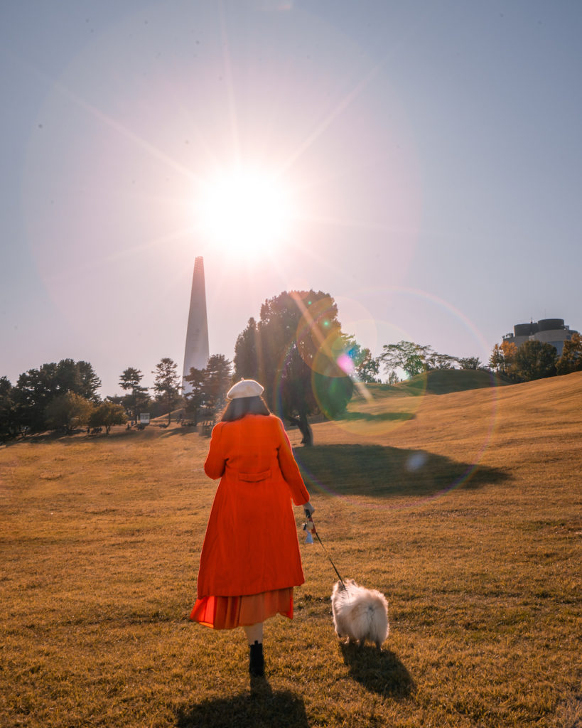 Woman in an orange coat walking a fluffy Pomeranian across a sunlit grassy hill at Olympic Park