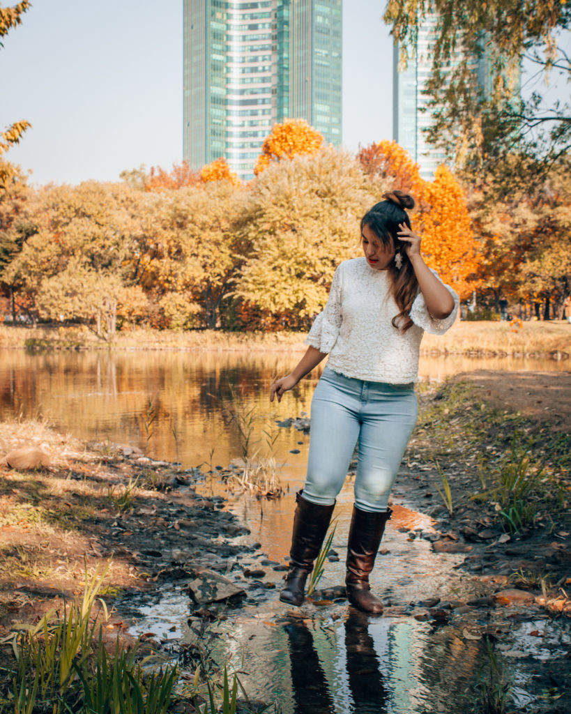 Woman in a white sweater and jeans standing in shallow water beside a pond at Seoul Forest, with golden autumn trees behind her